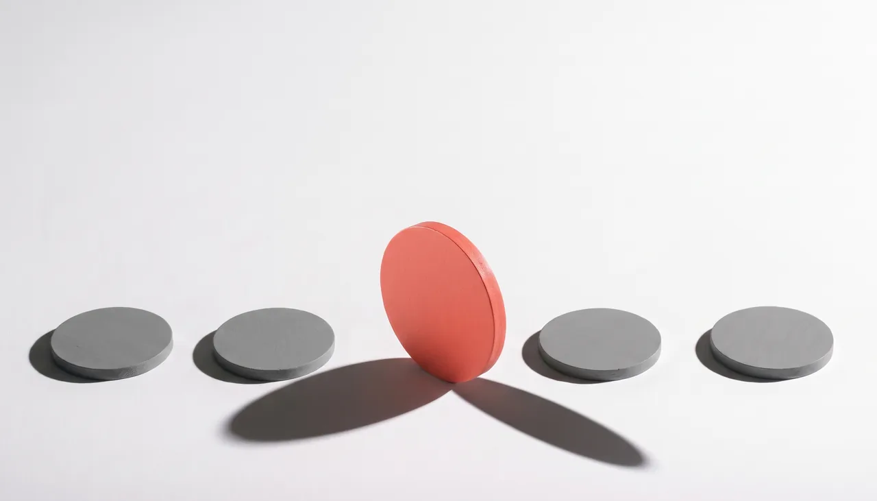 Conceptual still life: a row of identical flat grey discs on an off-white studio sweep, and one larger coral disc standing upright at the centre, casting a long hard shadow.